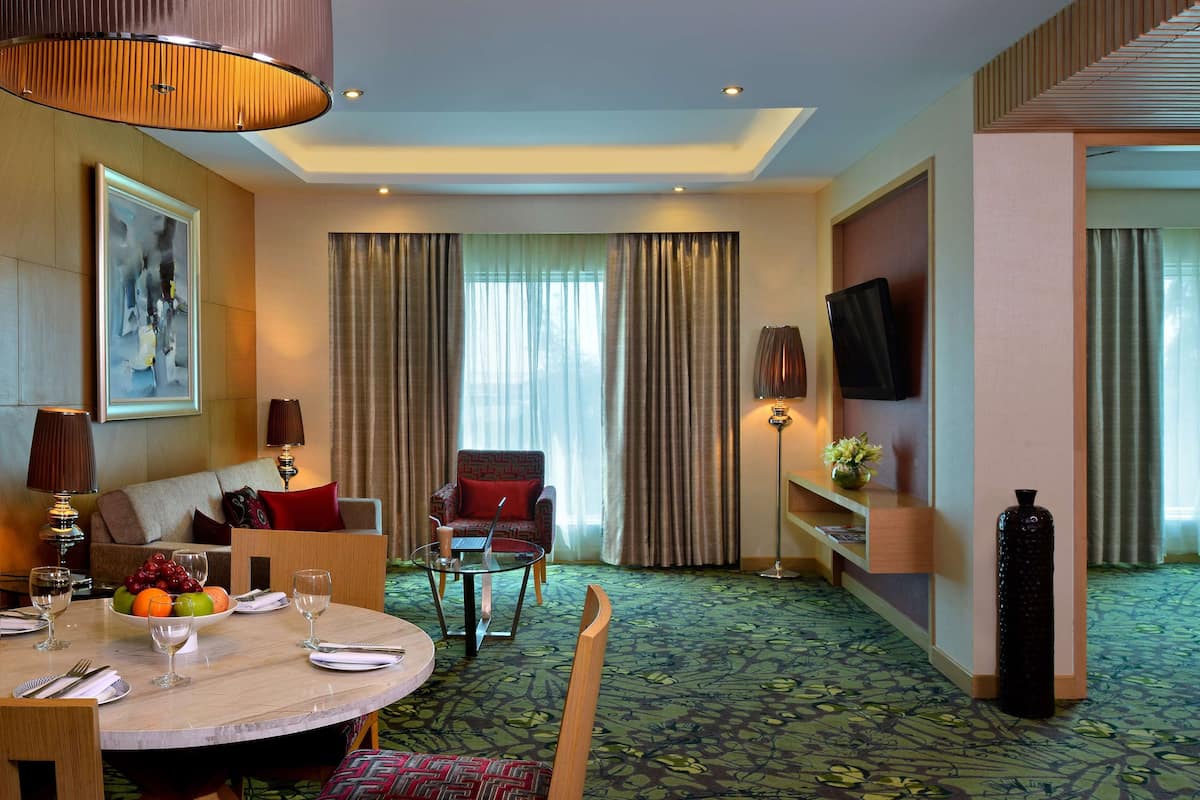 Habitación de hotel con comedor, sofá, sillón, televisión y un amplio ventanal con cortinas.