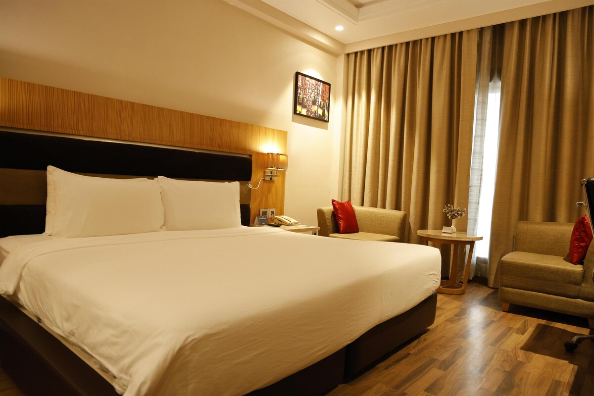 Camera d'albergo con un letto grande, una testiera in legno, un tavolino con una lampada, una sedia e vista all'esterno attraverso delle tende.