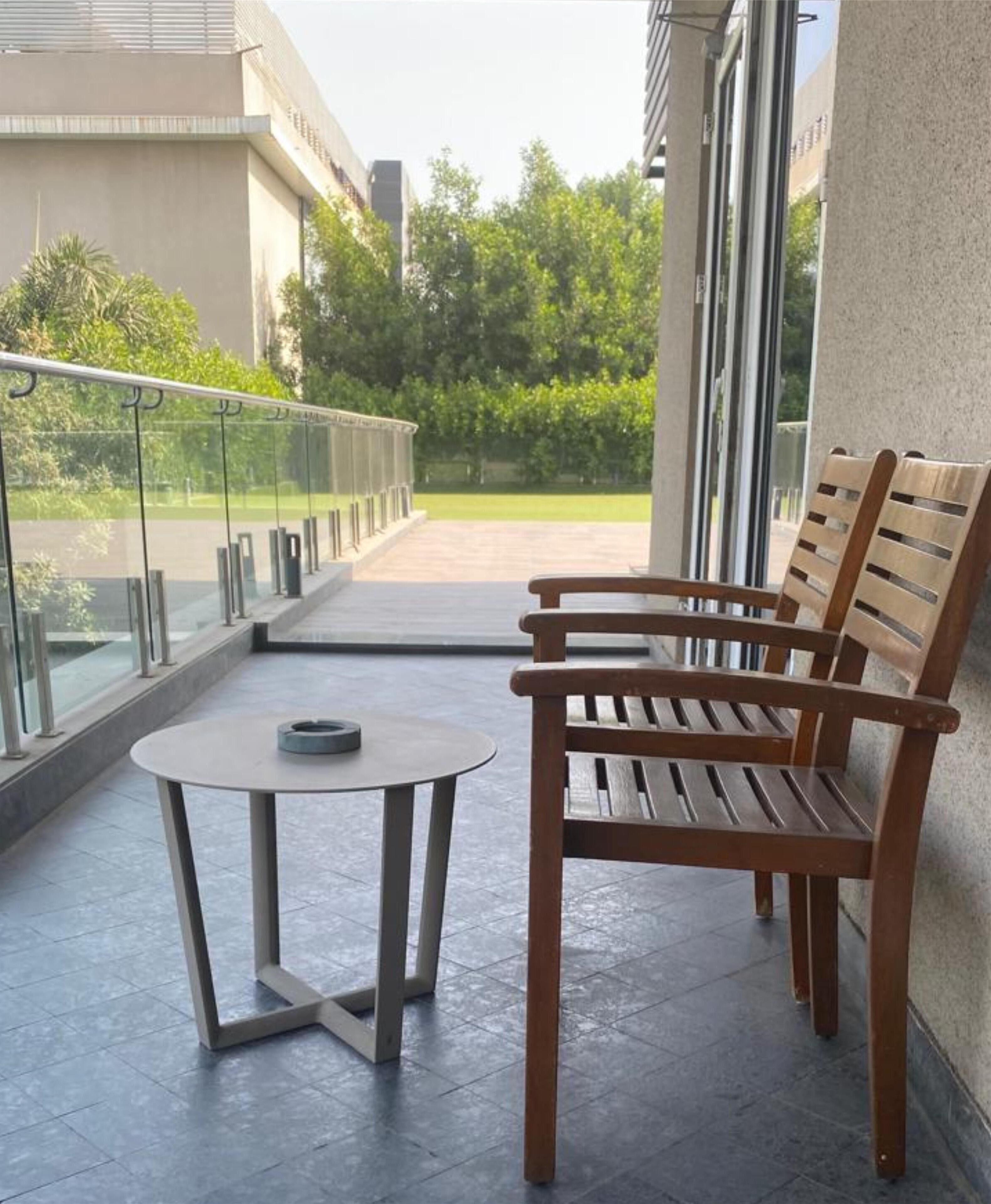 Un balcón con dos bancos de madera y una pequeña mesa redonda, con vista a un área verde con árboles y un edificio al fondo.