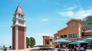 Una plaza rectangular con una torre del reloj, un edificio con balcón y mesas exteriores con sombrillas.