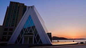 Un edificio moderno con fachada triangular y grandes ventanales, situado cerca de la costa con vistas al atardecer.