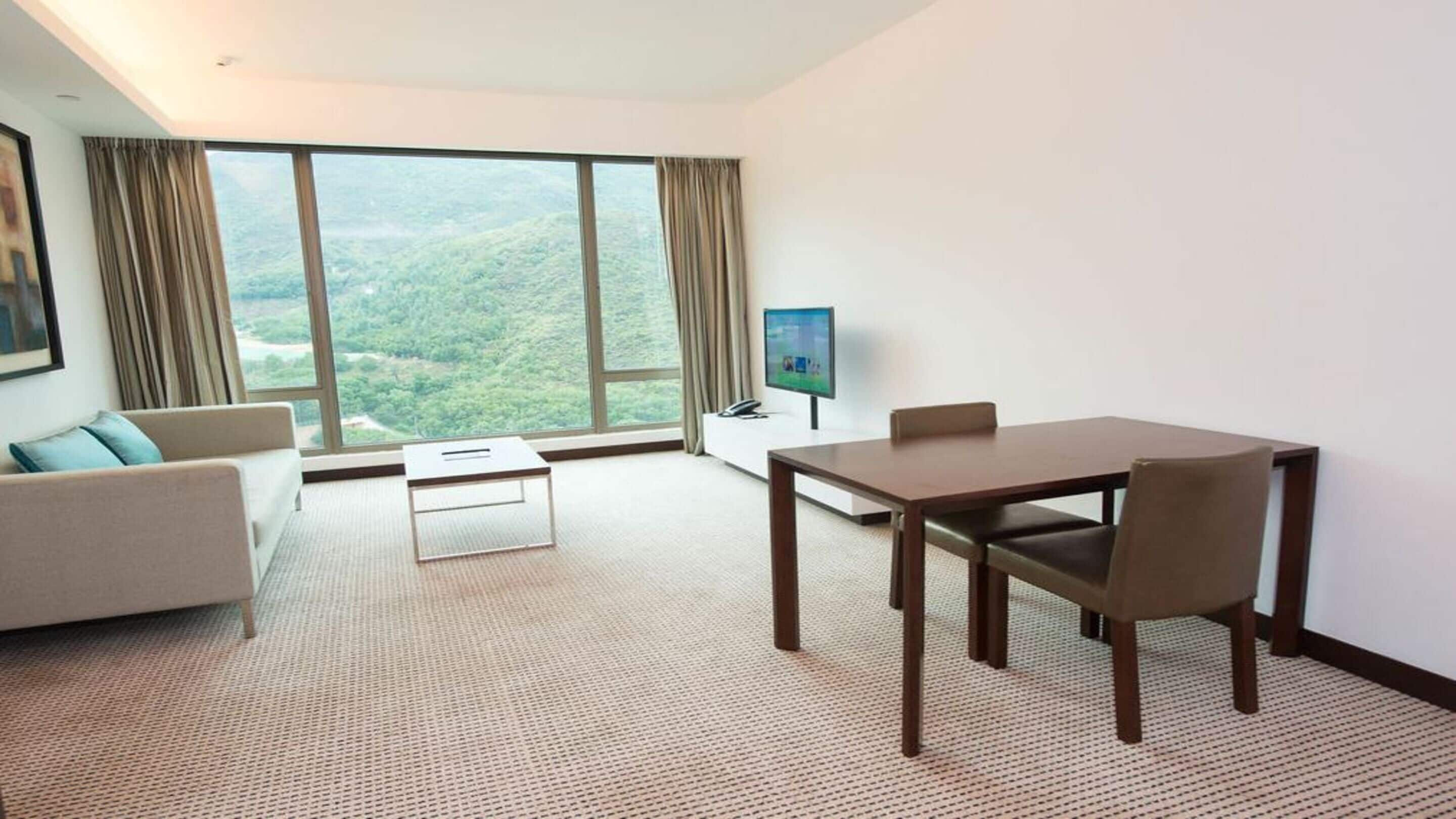 A hotel room with a large window, a sofa, a dining table, and a TV.