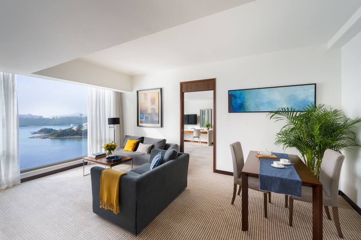 A modern hotel room with a large window offering a city view, a cozy seating area with a sofa and coffee table, a dining table with chairs, and a potted plant.