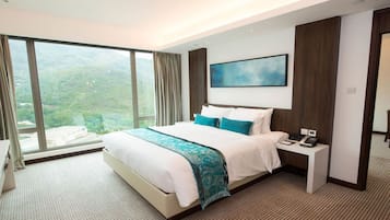 A modern hotel room with a large bed, a bedside table with a phone, and a view of a mountainous landscape through large windows.