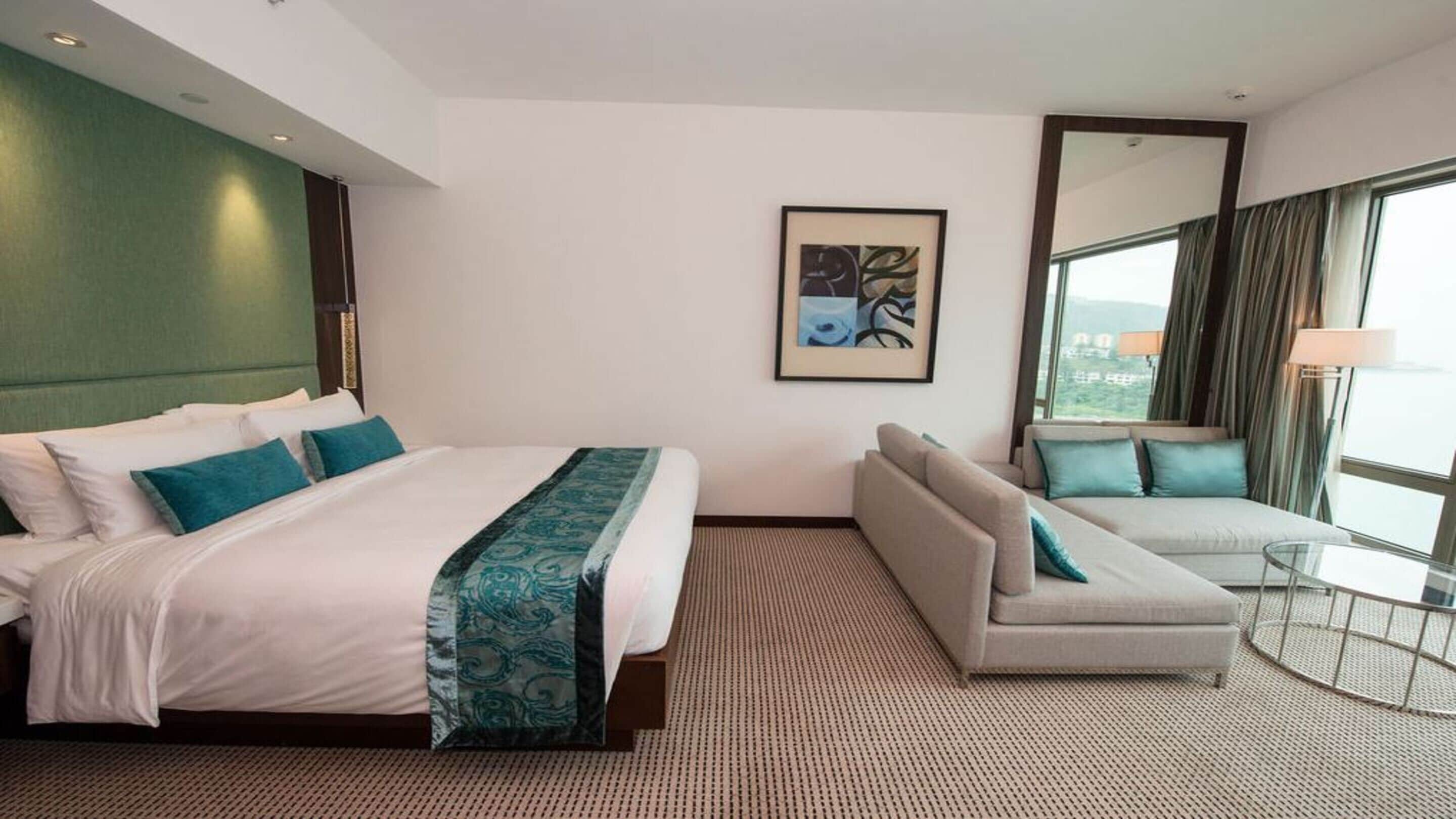 A hotel room with a large bed, a sofa, a small table, a mirror, and a window with a view.