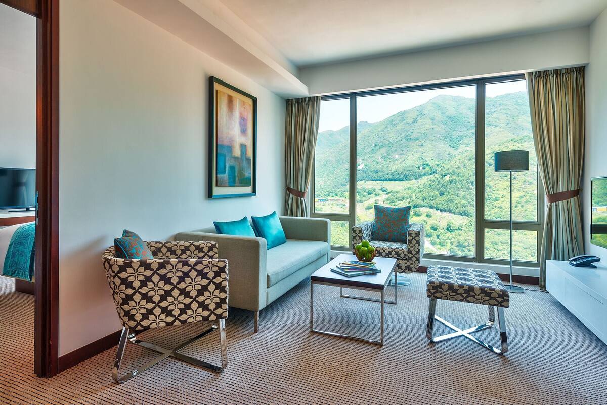A modern hotel room with a sofa, two armchairs, a coffee table, a TV, and a large window offering a mountain view.