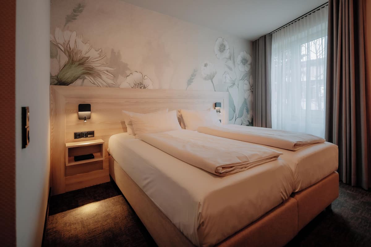 A hotel room with a large bed, a nightstand, a wall-mounted lamp, and a floral mural.