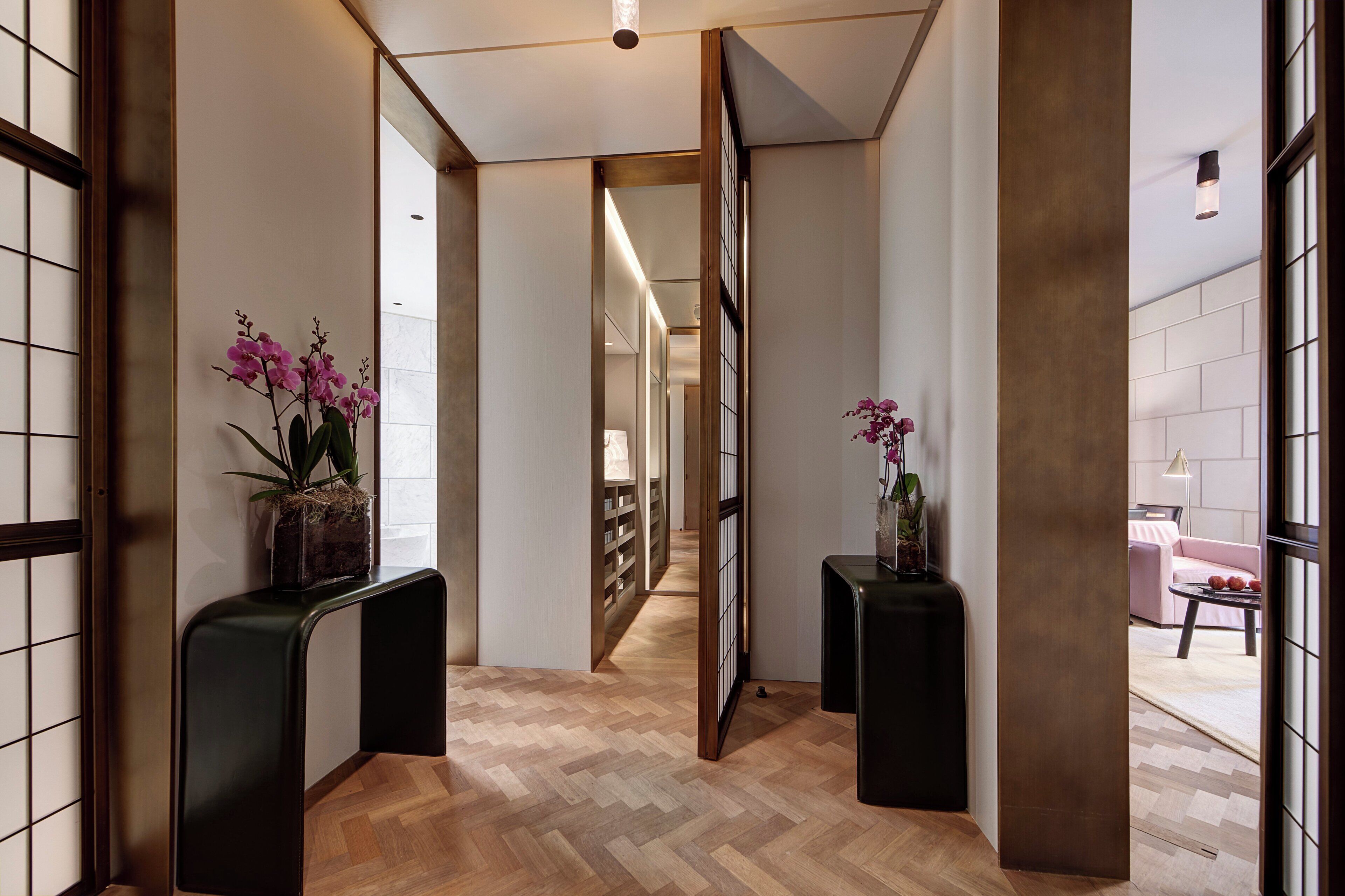 A hallway with wooden flooring, two black console tables with potted orchids, and a view into a room with a pink sofa and a table.