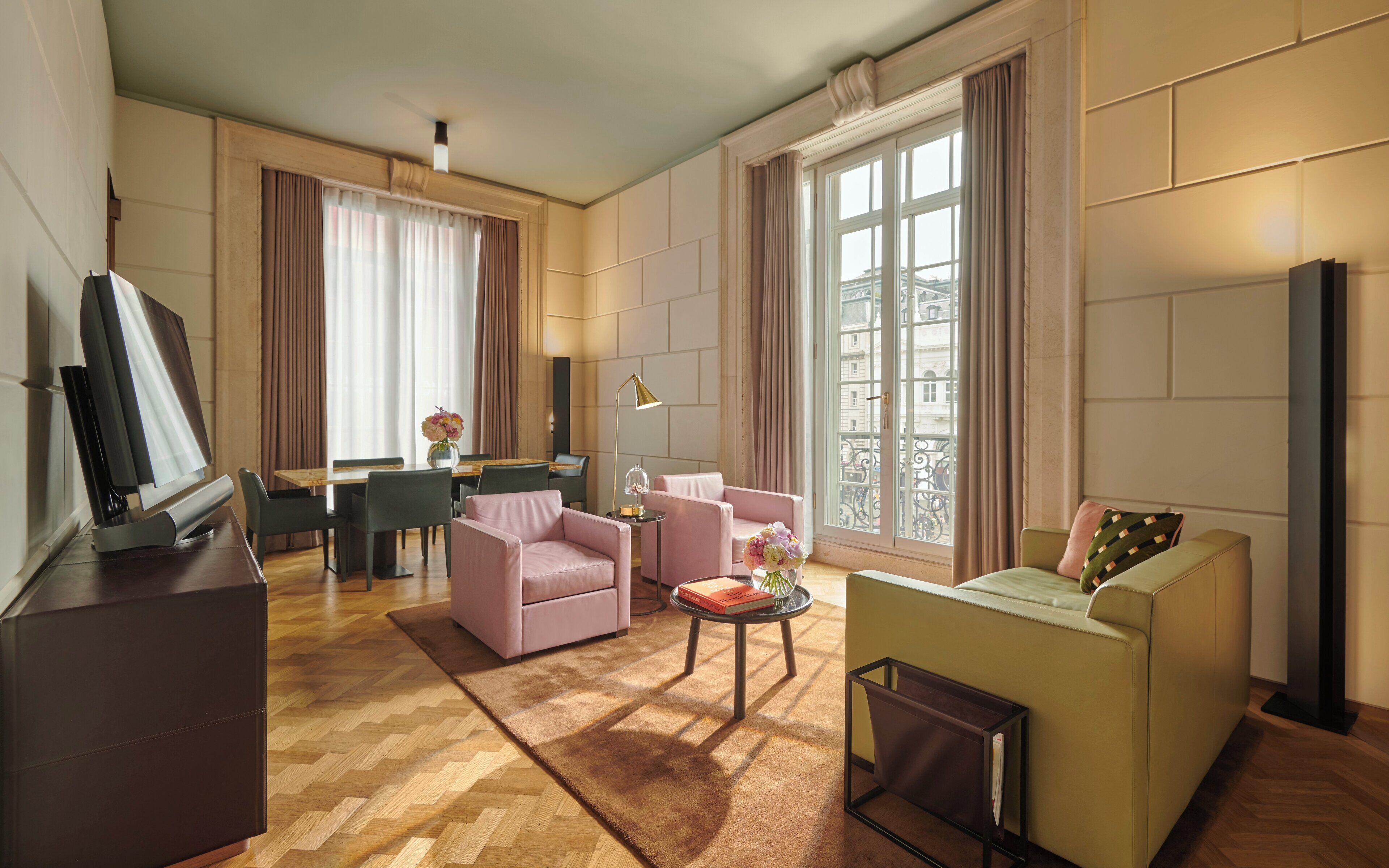 A modern living room with a flat-screen TV, a dining area with a table and chairs, and a large window with curtains.