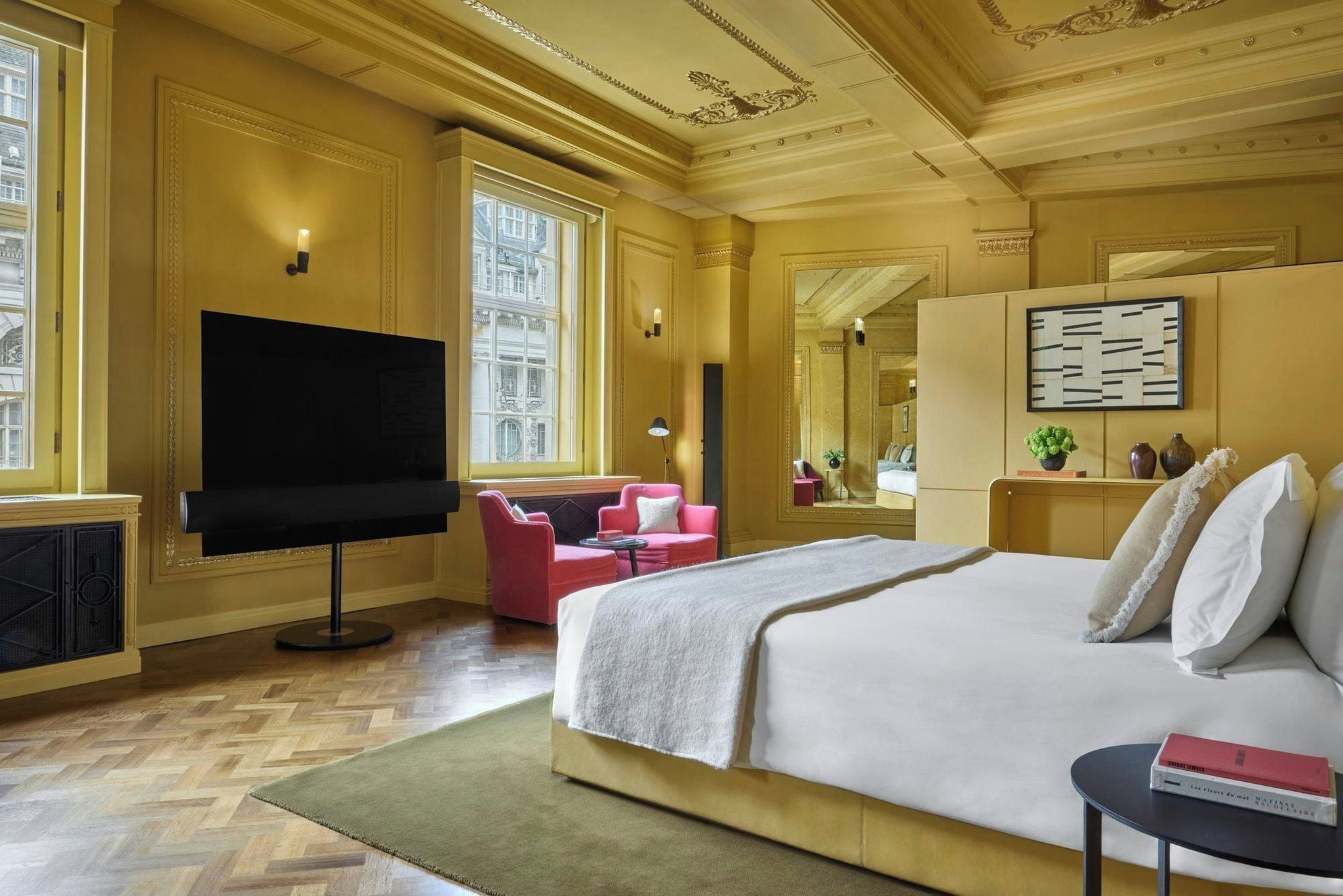 A hotel room with a large bed, a flat-screen TV, a pink armchair, a small table, and a large mirror.