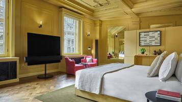 A hotel room with a large bed, a flat-screen TV, a pink armchair, a small table, and a large mirror.