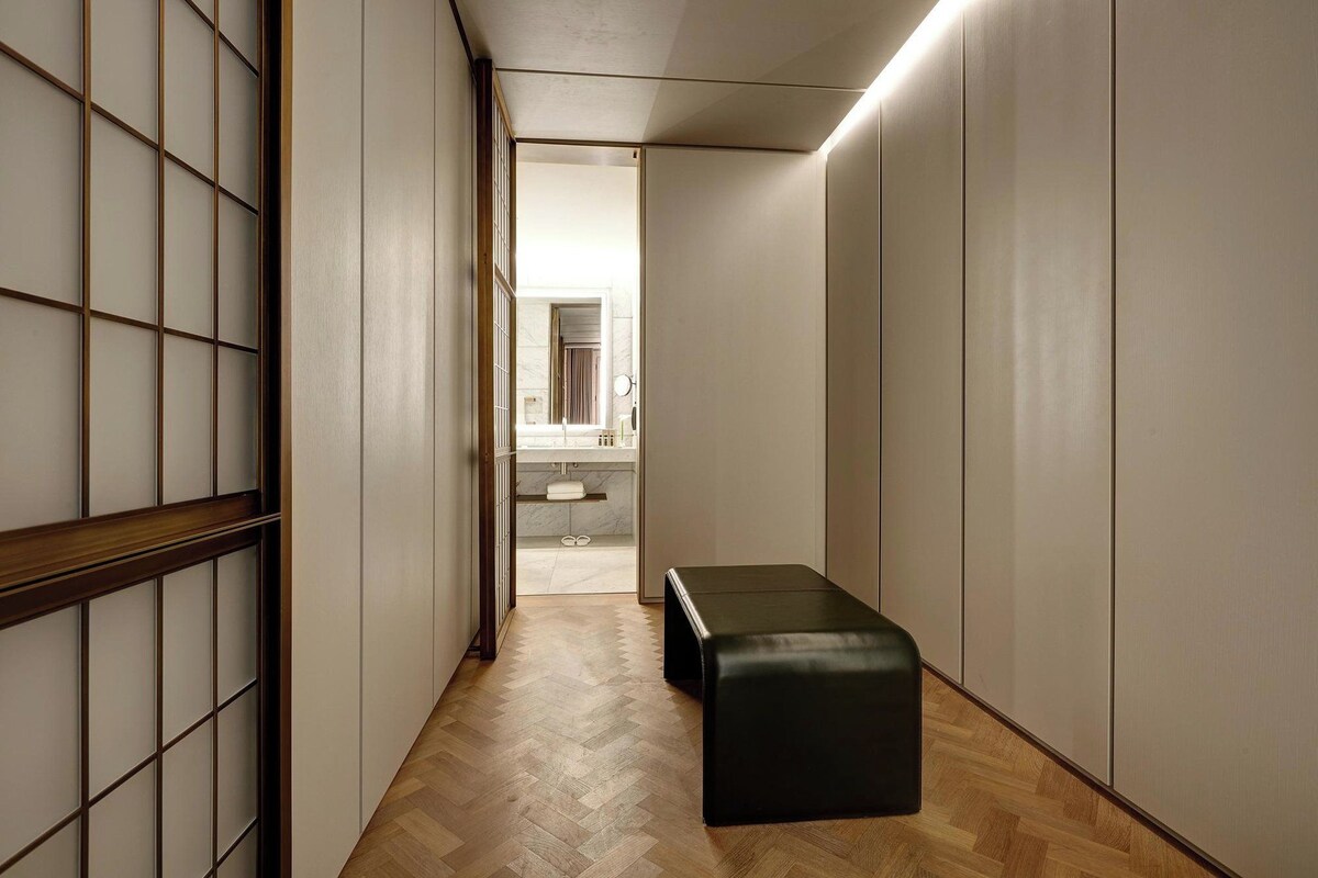 A hallway with a wooden floor, a bench, and a view into a bathroom with a mirror and a sink.