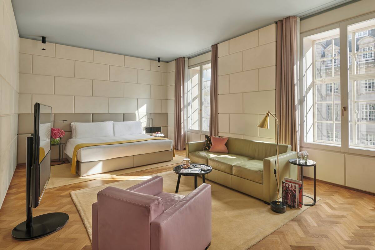 A modern hotel room with a bed, sofa, armchair, and a small table with a lamp.