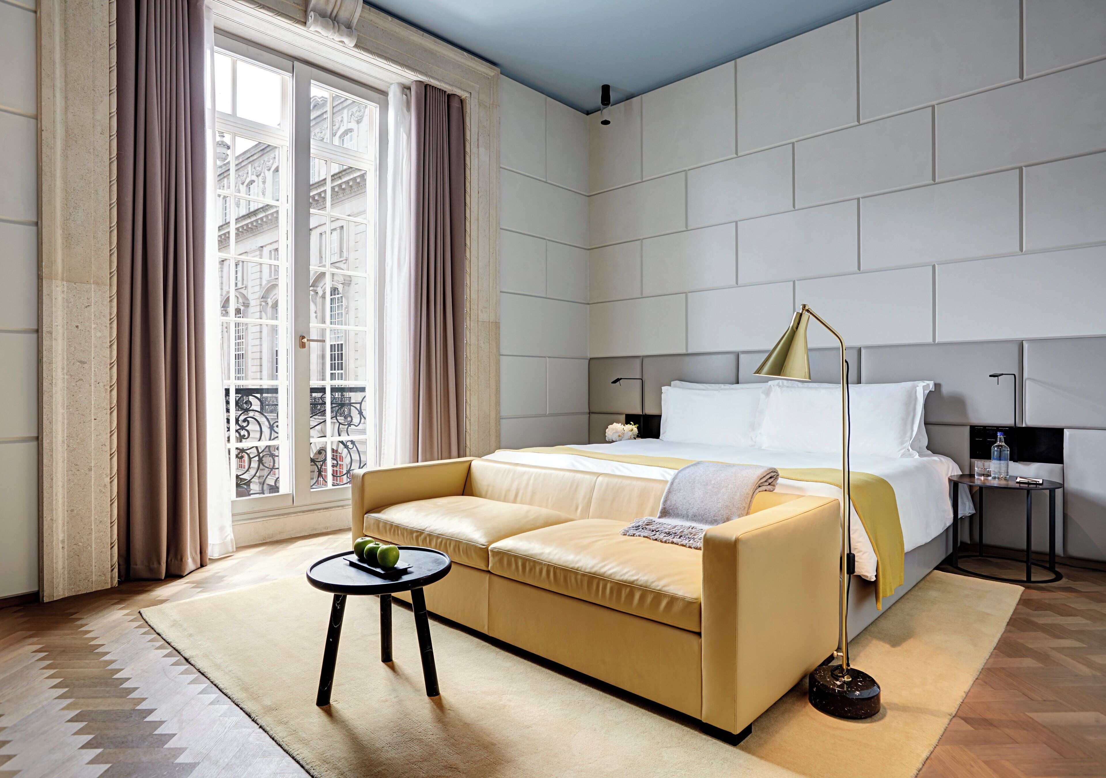 A hotel room with a large bed, a sofa, a small table with a plant, and a view of the city through large windows.