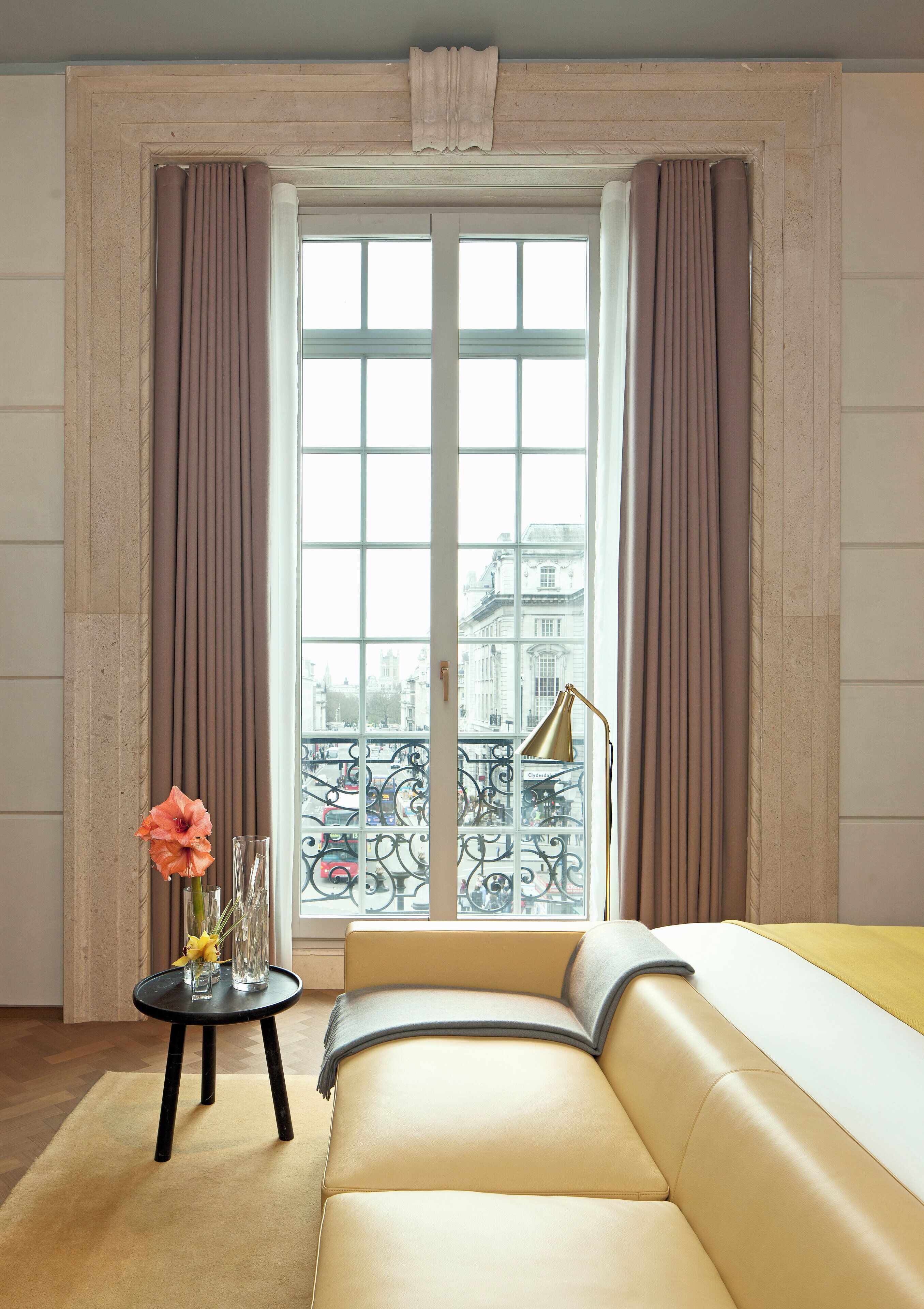 Een kamer met een bank, een klein tafeltje met een vaas, en een uitzicht op een stadsbeeld door een raam met gordijnen.