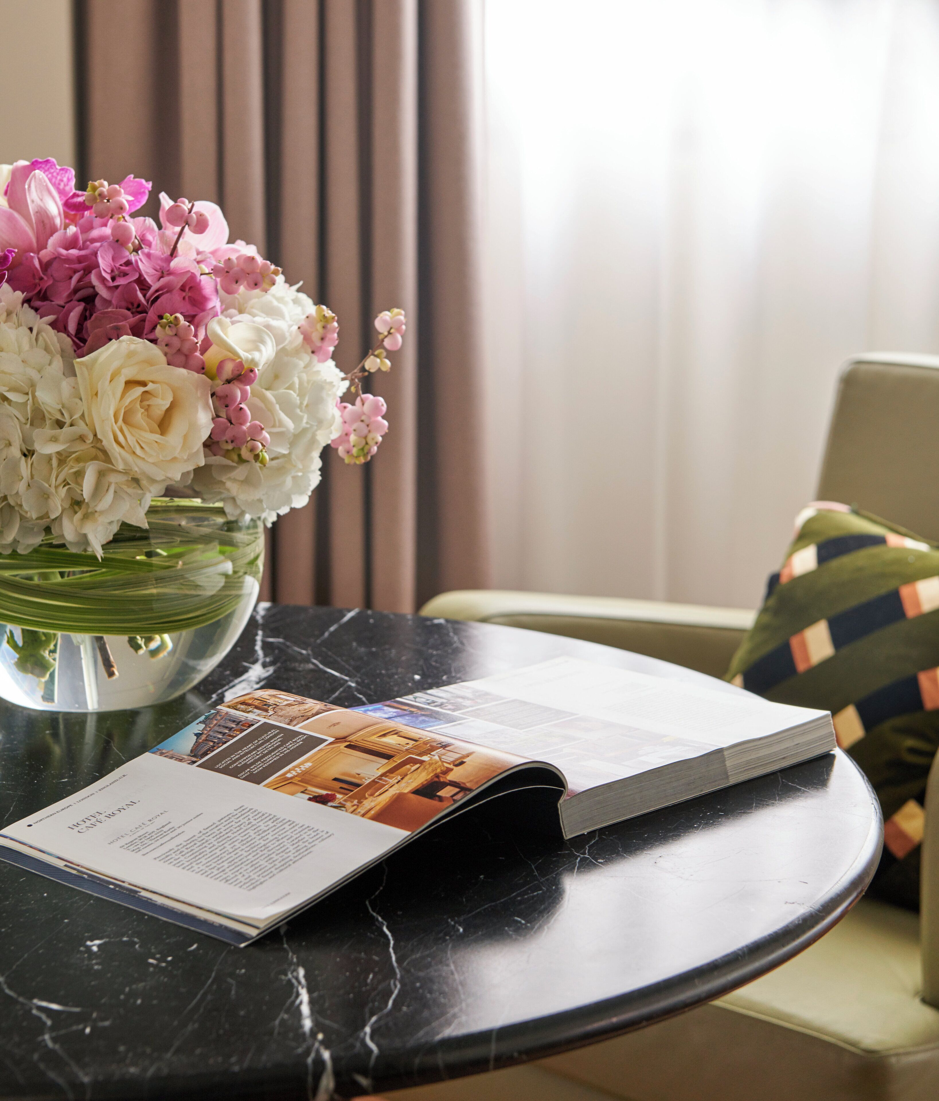 A magazine on a marble table with a floral arrangement in a clear vase.