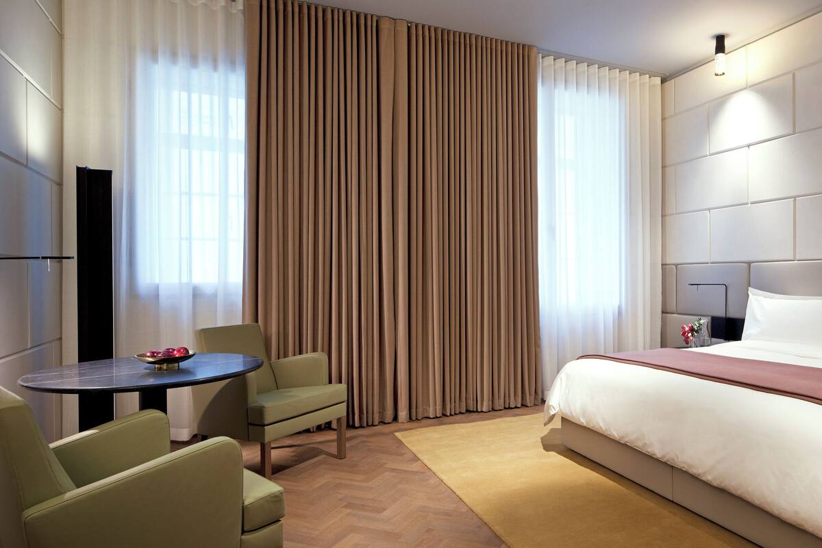 A hotel room with a bed, a desk, two chairs, and a large window with curtains.
