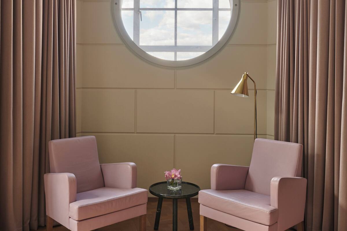 A room with two pink armchairs, a small round table with a vase of flowers, and a large window with curtains.