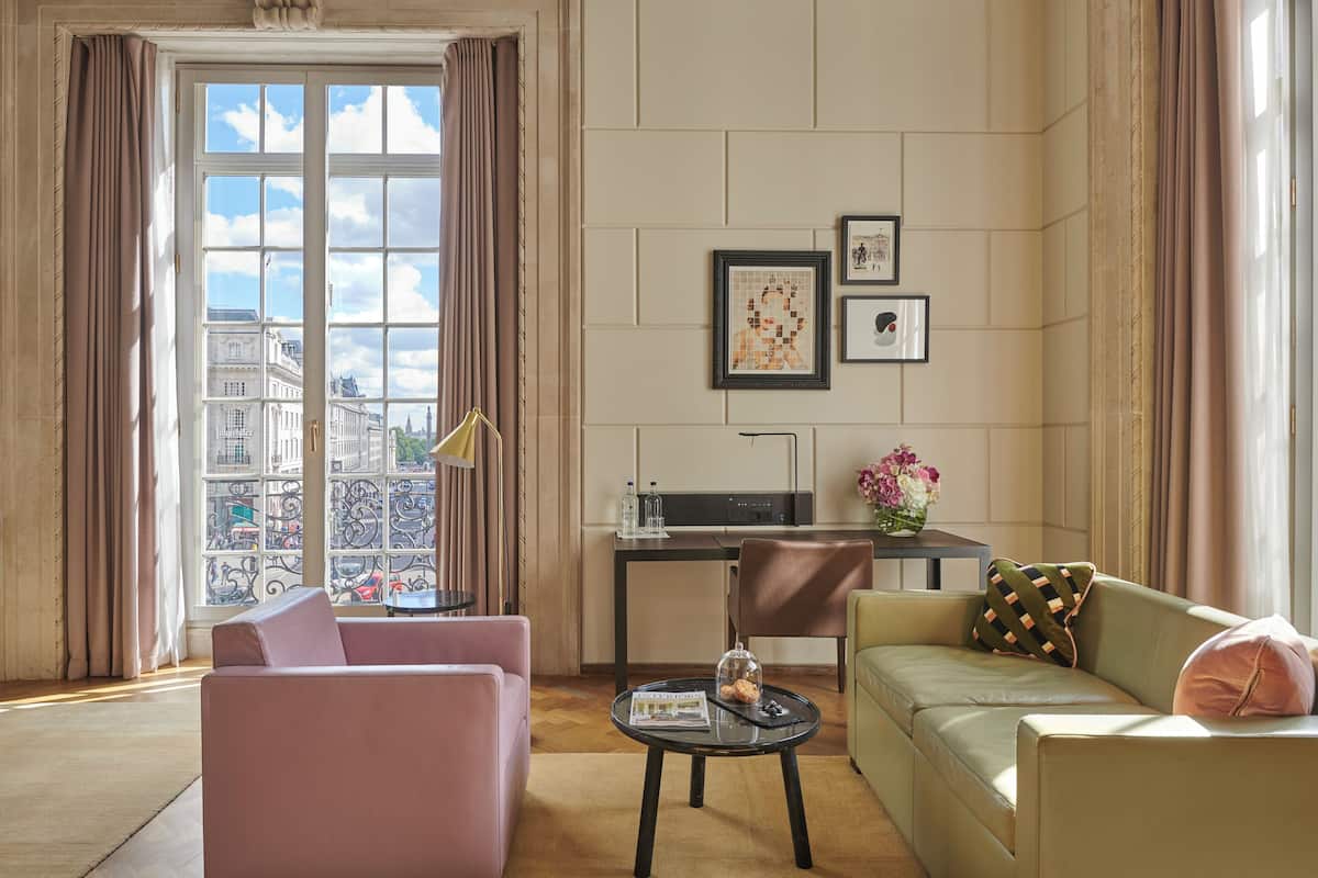 A hotel room with a large window, a sofa, a dining table, and a small kitchen area.