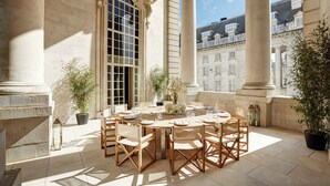 An outdoor dining area with a round table set for six, surrounded by wooden chairs. There are potted plants and a large arched window. - Hotel Cafe Royal, London (London)