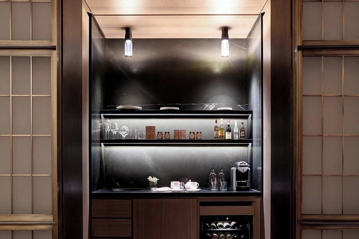 A modern, dark wood-paneled bar area with overhead lighting, shelves of glassware, bottles, and a wine cooler.