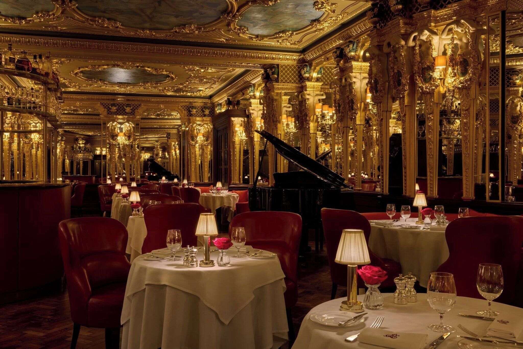 A luxurious dining room with ornate gold detailing, multiple tables set for dining, and a grand piano.