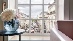 A view through a window of a busy street with historic buildings, a vase with flowers on a table, and a pink sofa. - Hotel Cafe Royal, London (London)