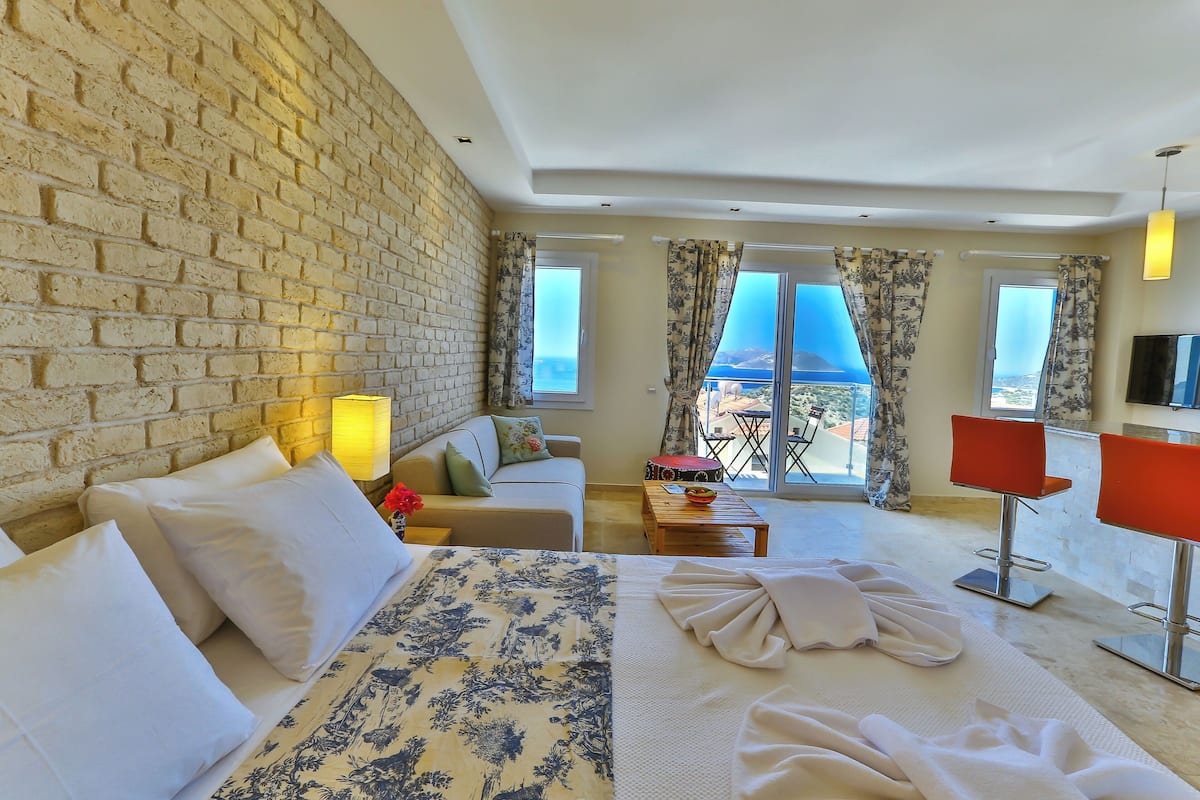 A hotel room with a brick wall, a bed with a floral bedspread, a sofa, a coffee table, a TV, and a balcony with a view.