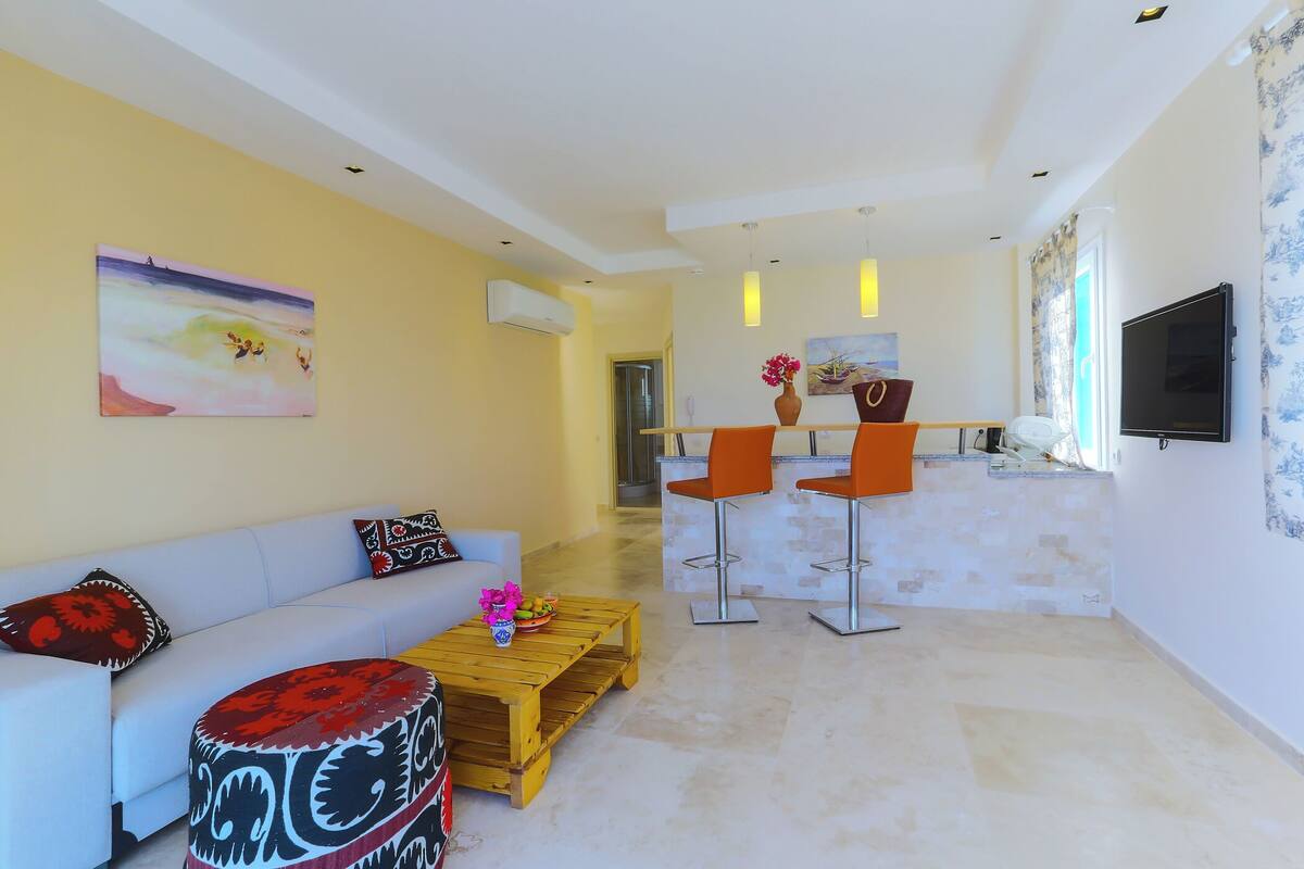 A modern living room with a sofa, coffee table, and two orange chairs. A flat-screen TV is mounted on the wall, and there is a painting above the sofa.