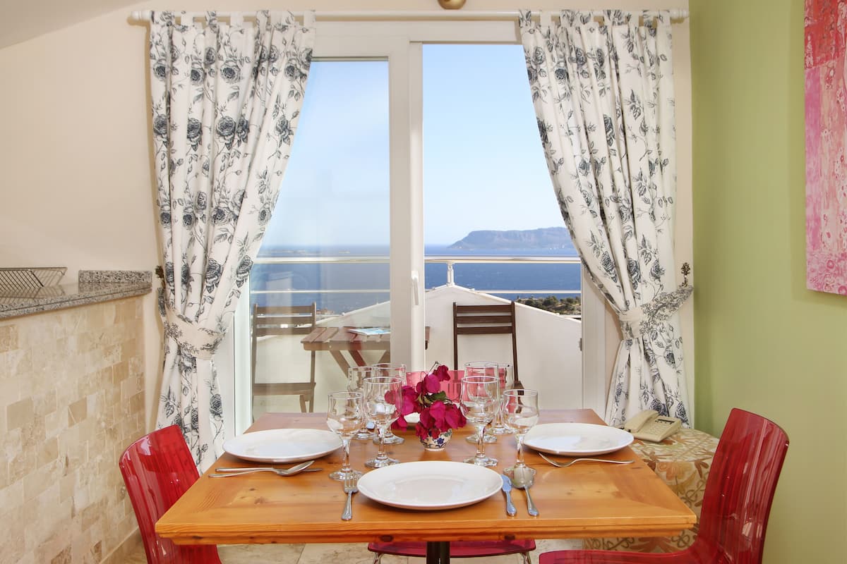 A dining table set with plates, glasses, and cutlery, with a view of the sea and mountains through a window.