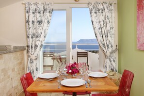 Una mesa de comedor dispuesta con platos, vasos y cubiertos, con vista al mar y a las montañas a través de una ventana.