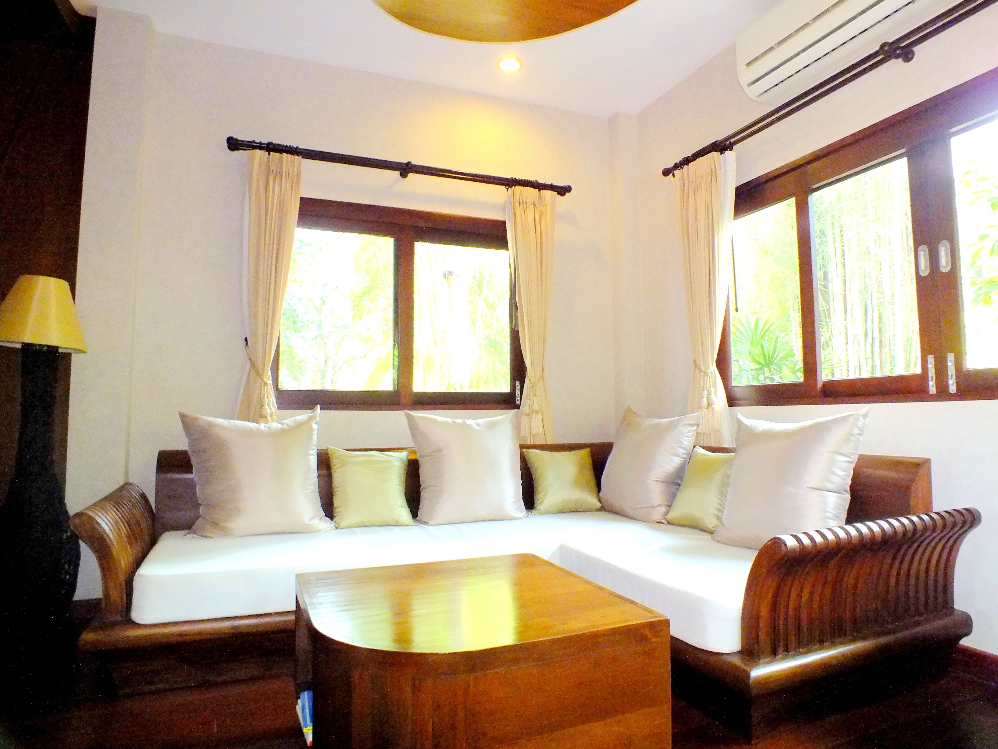A room with a wooden sofa, a wooden coffee table, and a lamp. There are windows with curtains and a view of greenery outside.