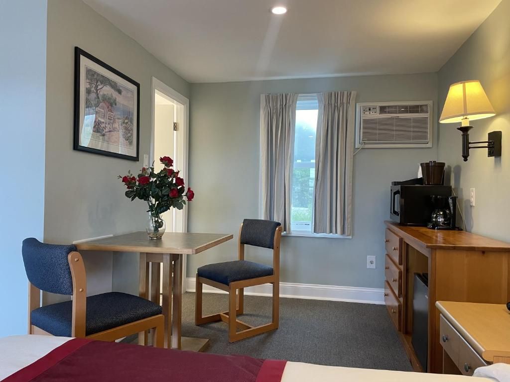 A hotel room with a bed, a desk, a chair, a vase with flowers, a window with curtains, and a lamp.