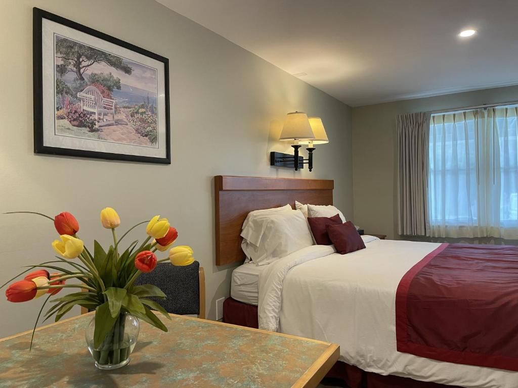 A hotel room with a bed, a nightstand, a lamp, a framed picture on the wall, and a vase with flowers on a table.