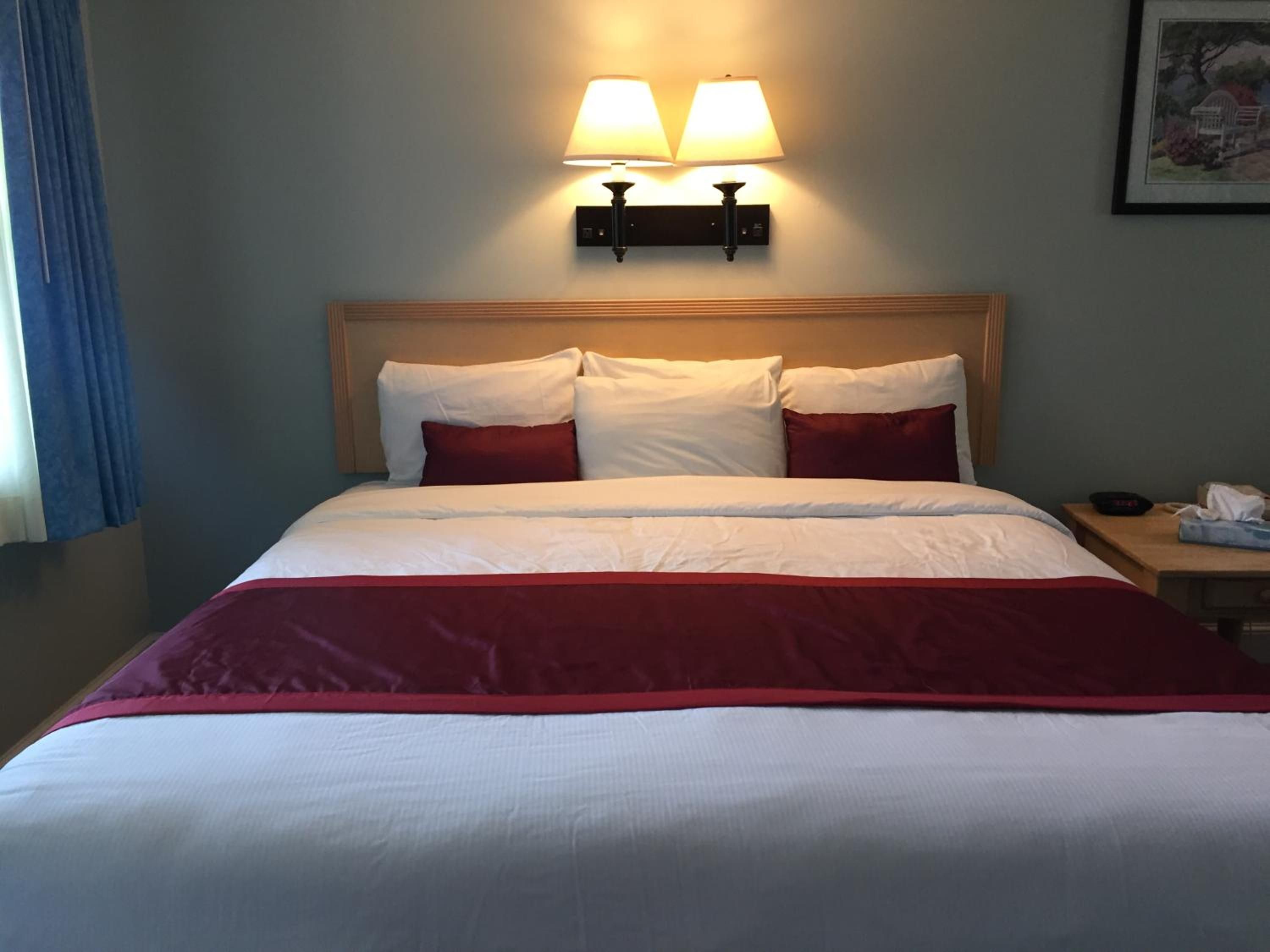 Un lit bien fait, avec une literie blanche et rouge, deux lampes fixées au mur et un tableau encadré accroché au mur.