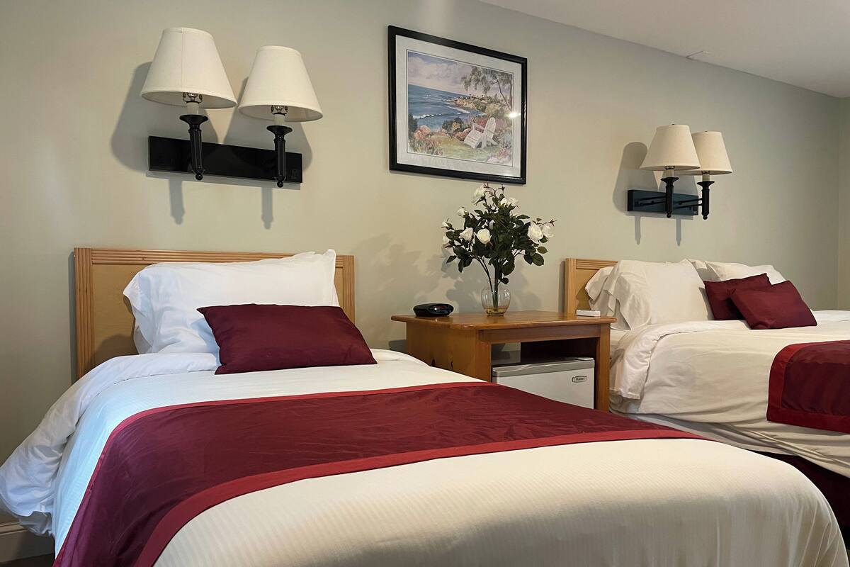 A hotel room with two beds, each with white linens and red accents, a nightstand with a lamp, a framed painting of a coastal scene, and a small vase with flowers.