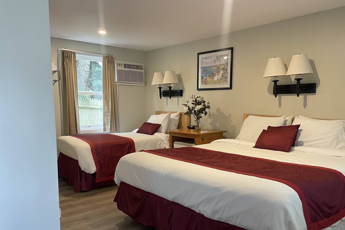 A hotel room with two beds, beige walls, a window with curtains, a painting, and two wall-mounted lamps.