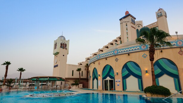 Um hotel com piscina, palmeiras e um edifício grande com janelas em arco e uma torre.