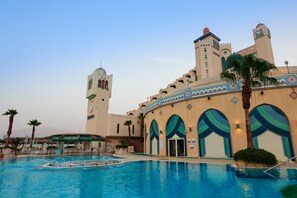 Un hôtel doté d’une piscine, de palmiers et d’un grand bâtiment aux fenêtres en arc de cercle et surmonté d’une tour.