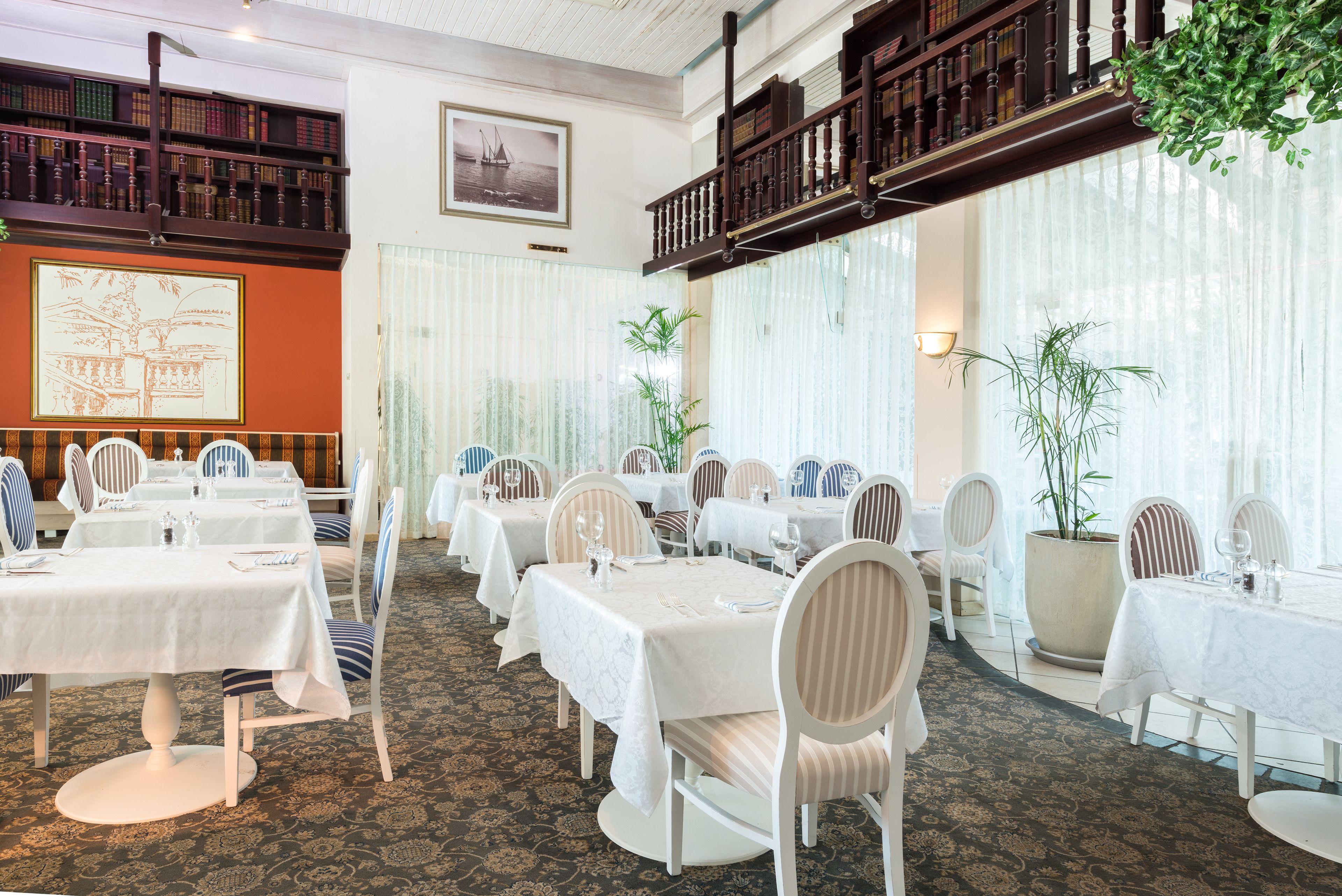 A formal dining room with multiple tables and chairs, a balcony with bookshelves, and large windows with sheer curtains.