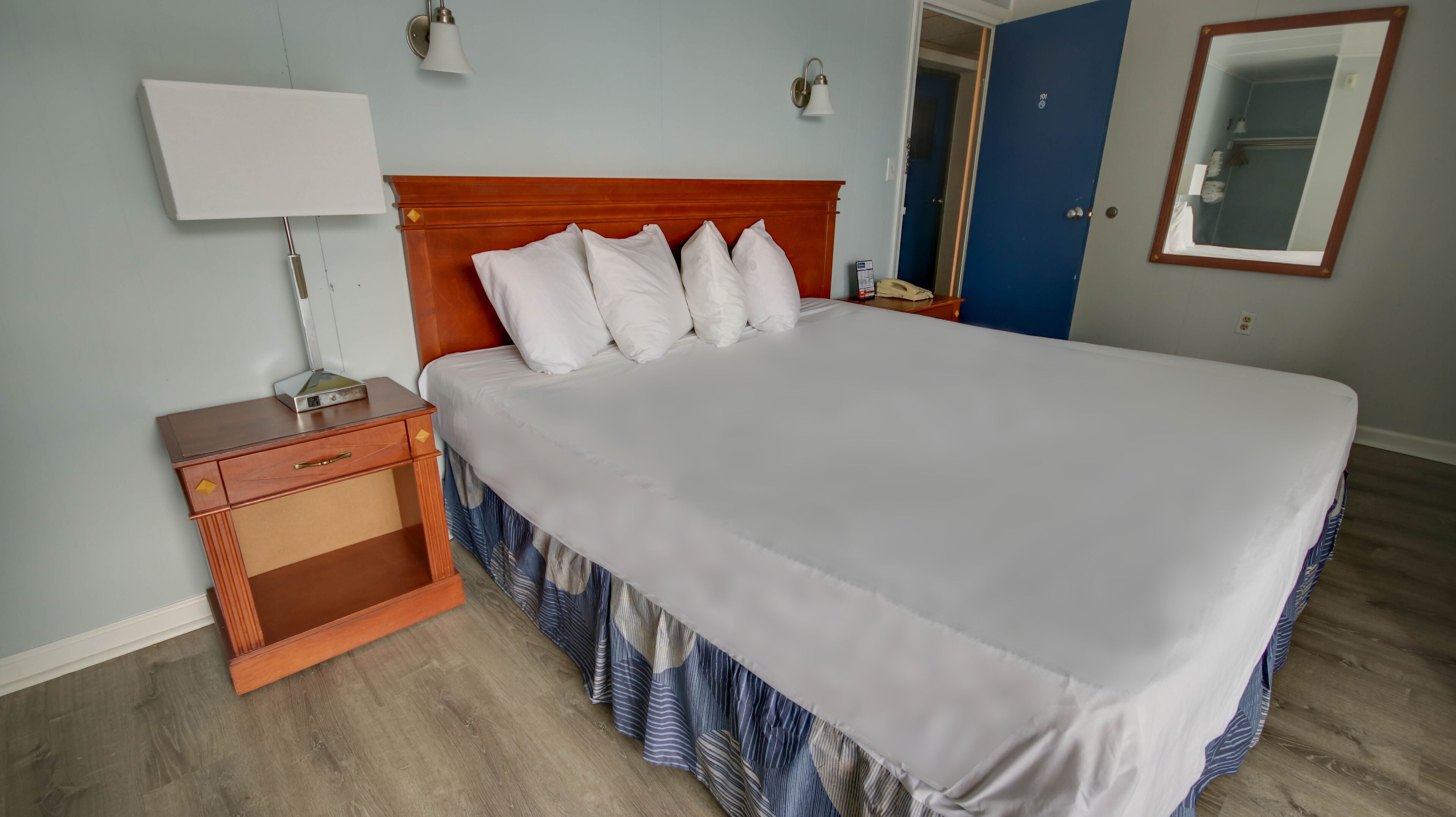 A neatly made bed with white linens and a wooden headboard, a bedside table with a lamp, and a blue door in the background.