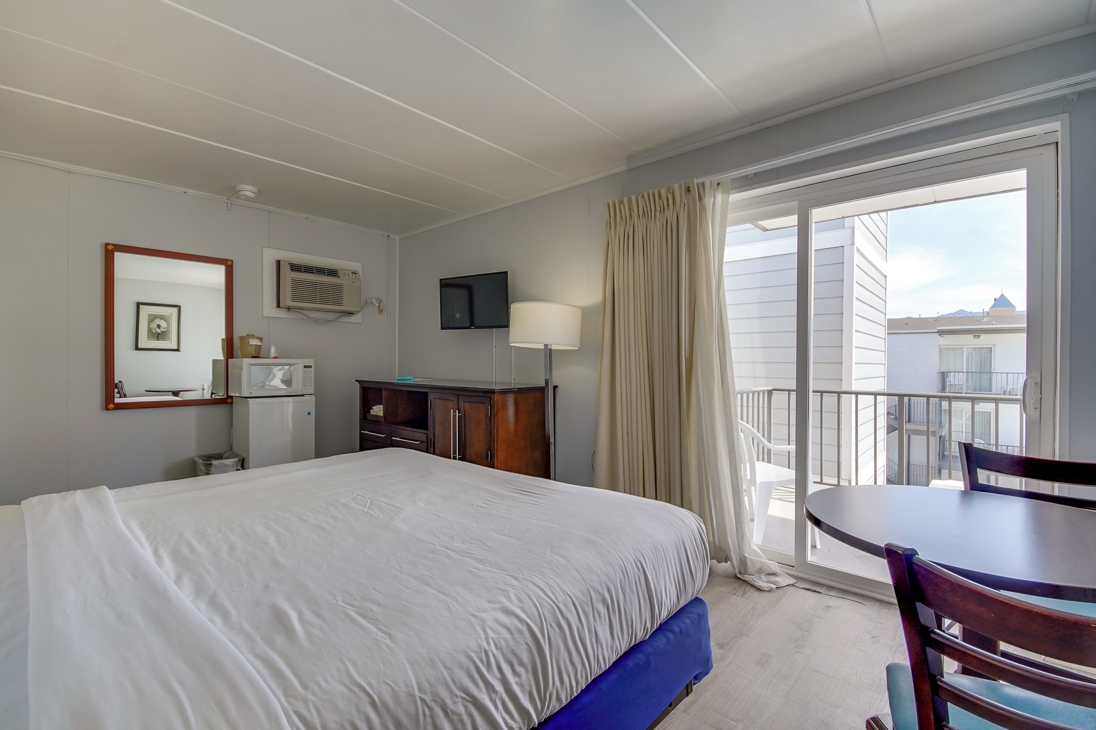 A hotel room with a bed, a desk with a chair, a TV, a microwave, and a balcony with a view of buildings.