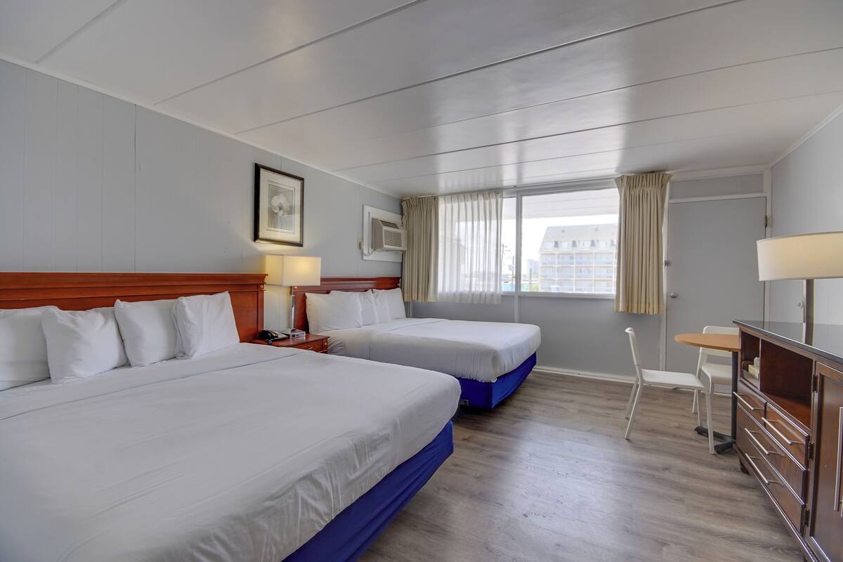 Une chambre d’hôtel avec deux lits, un bureau, une chaise, une lampe et une fenêtre avec des rideaux.