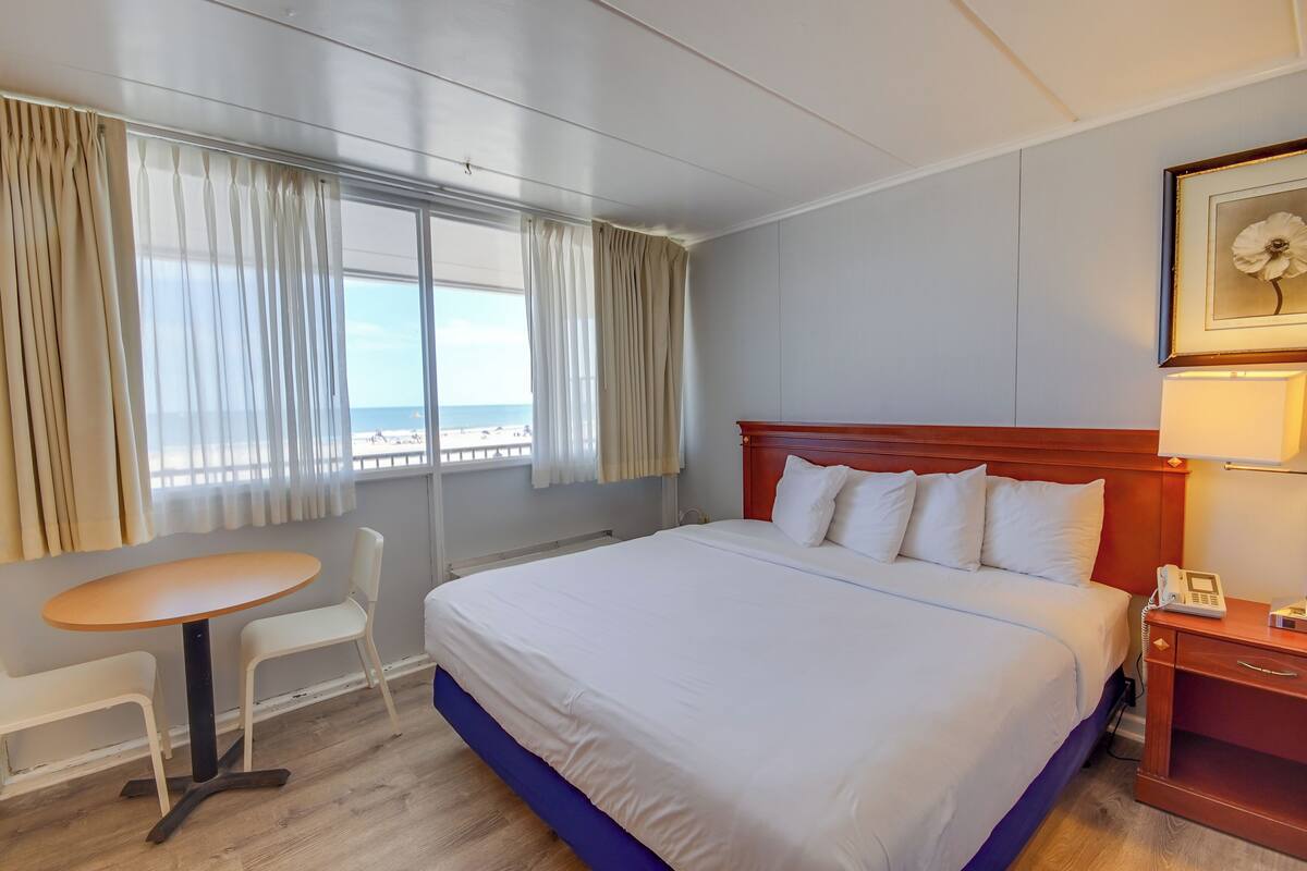 Une chambre d’hôtel avec un lit, une petite table avec des chaises, une table de chevet avec une lampe et une fenêtre avec des rideaux.