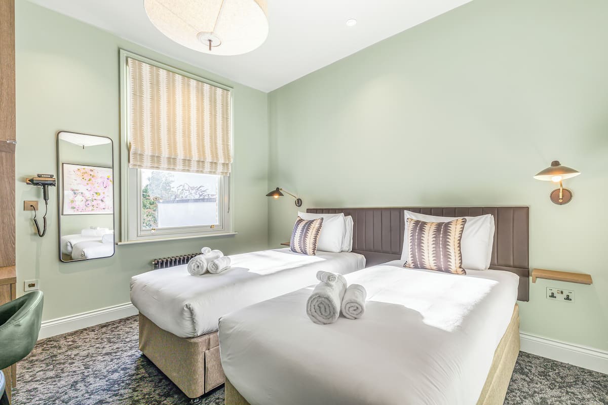 A hotel room with two beds, a large window with blinds, a mirror, a bedside table with a lamp, and a chair.