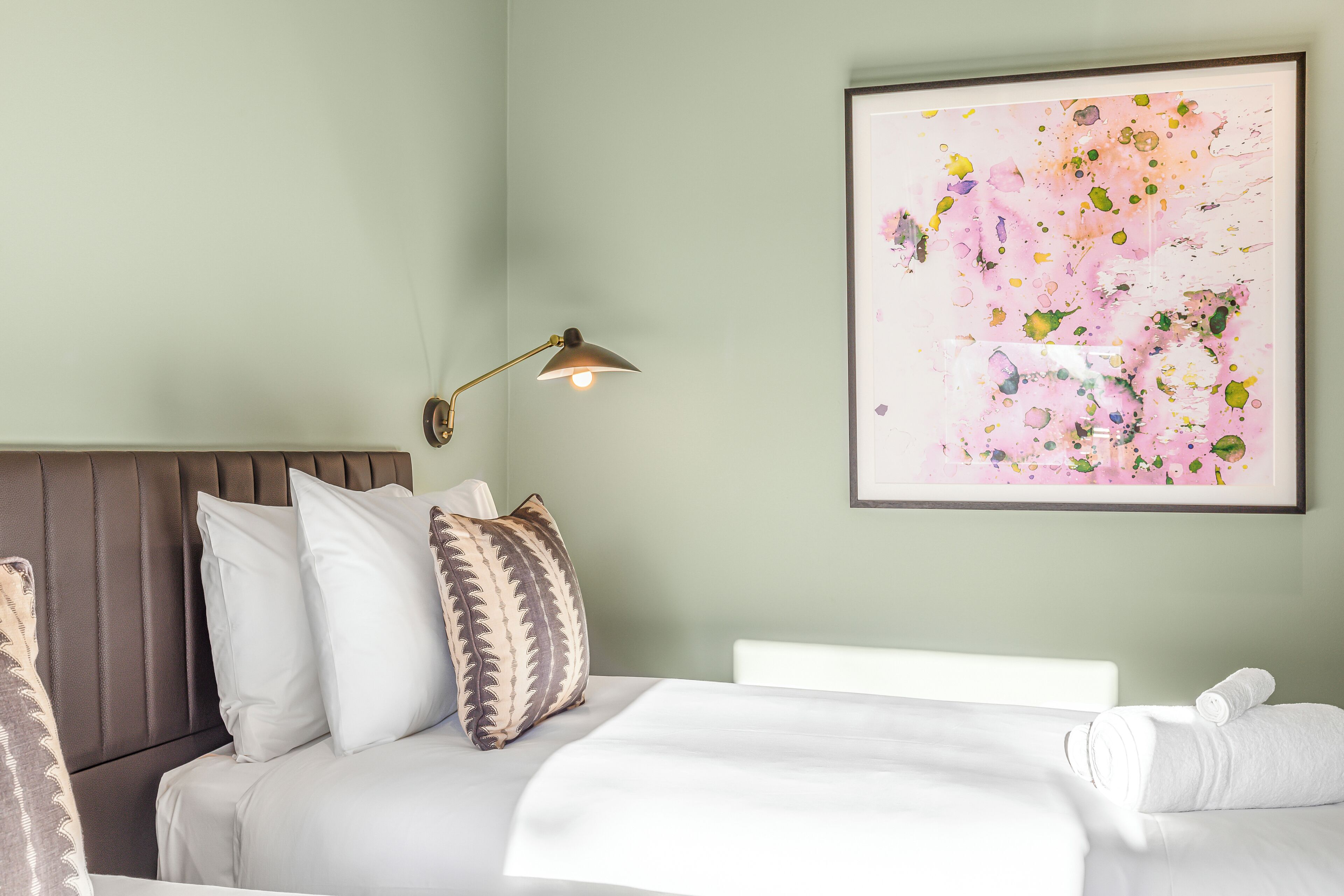A neatly made bed with white linens and a decorative pillow, a wall-mounted lamp, and a framed abstract painting.