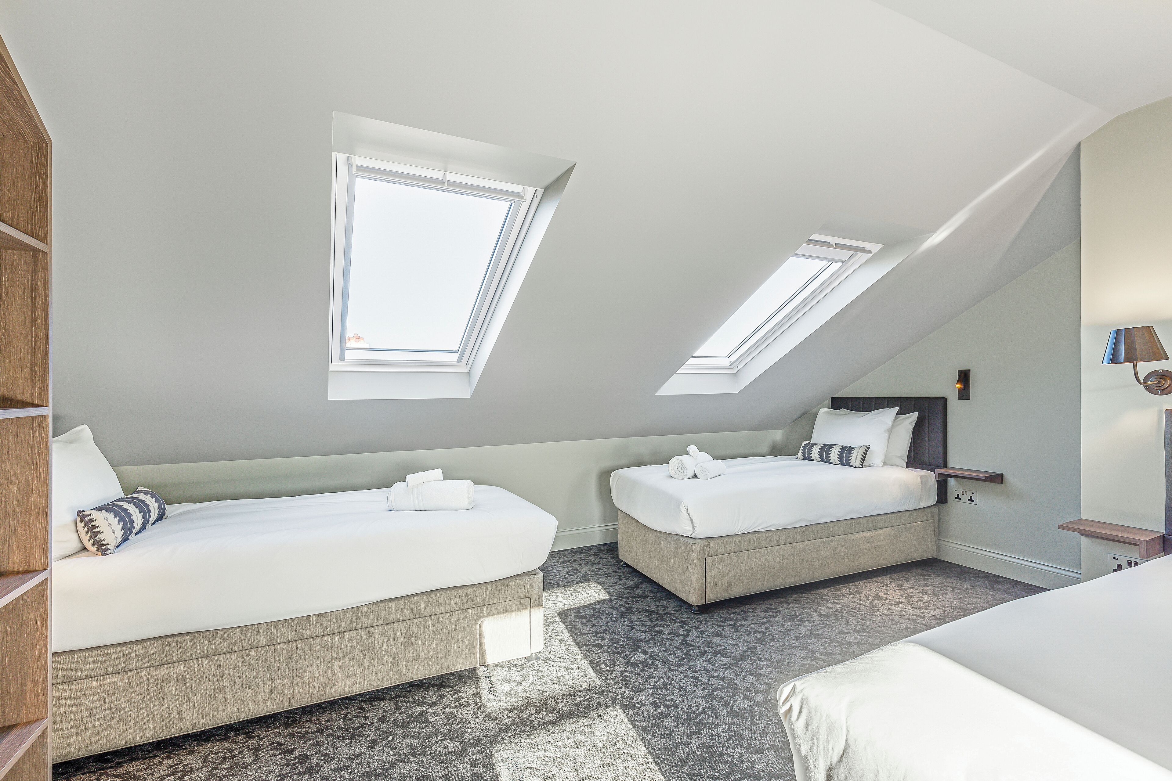 A modern hotel room with two beds, skylights, a wooden wardrobe, and a bedside table with a lamp.