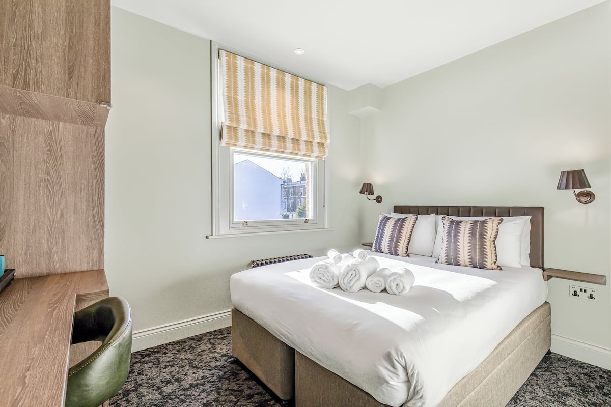 A hotel room with a bed, a desk, a chair, a window with a valance, and a view of buildings outside.