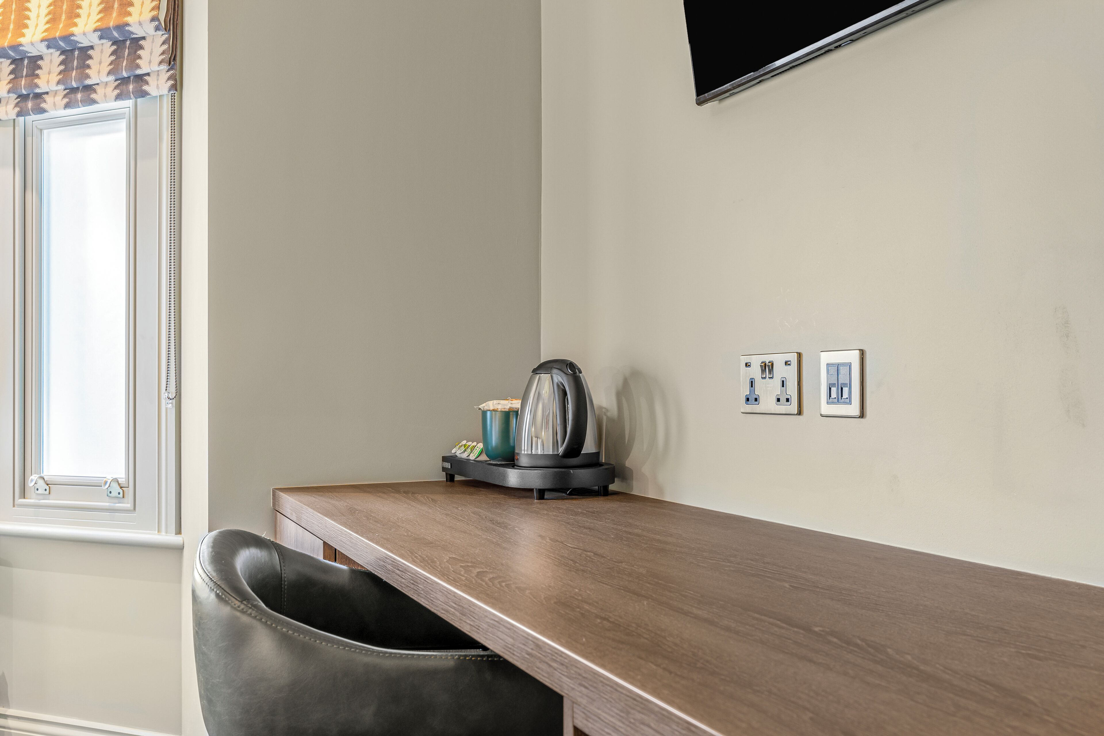 A room with a wooden desk, a black leather chair, a kettle, and a flat-screen TV mounted on the wall.