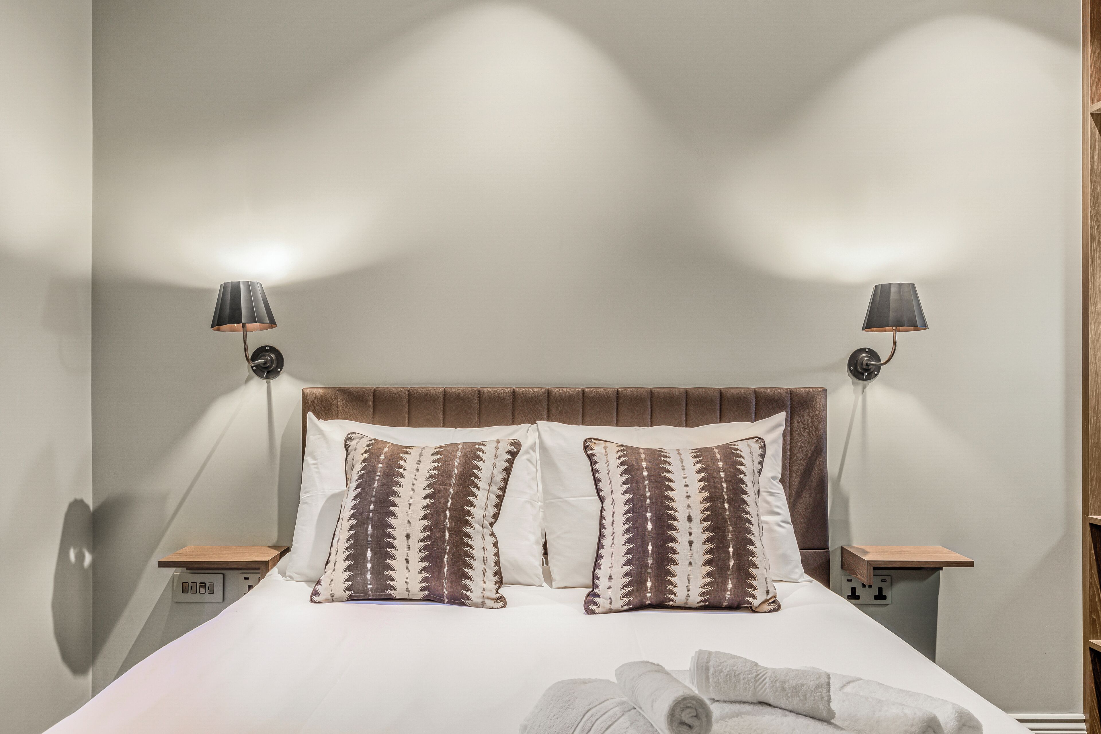 A neatly made bed with two pillows, a bedside table, and two wall-mounted lamps.